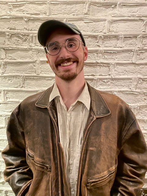 Mike Fox, smiling in a cap, round glasses, and a worn brown leather jacket against a white brick wall
