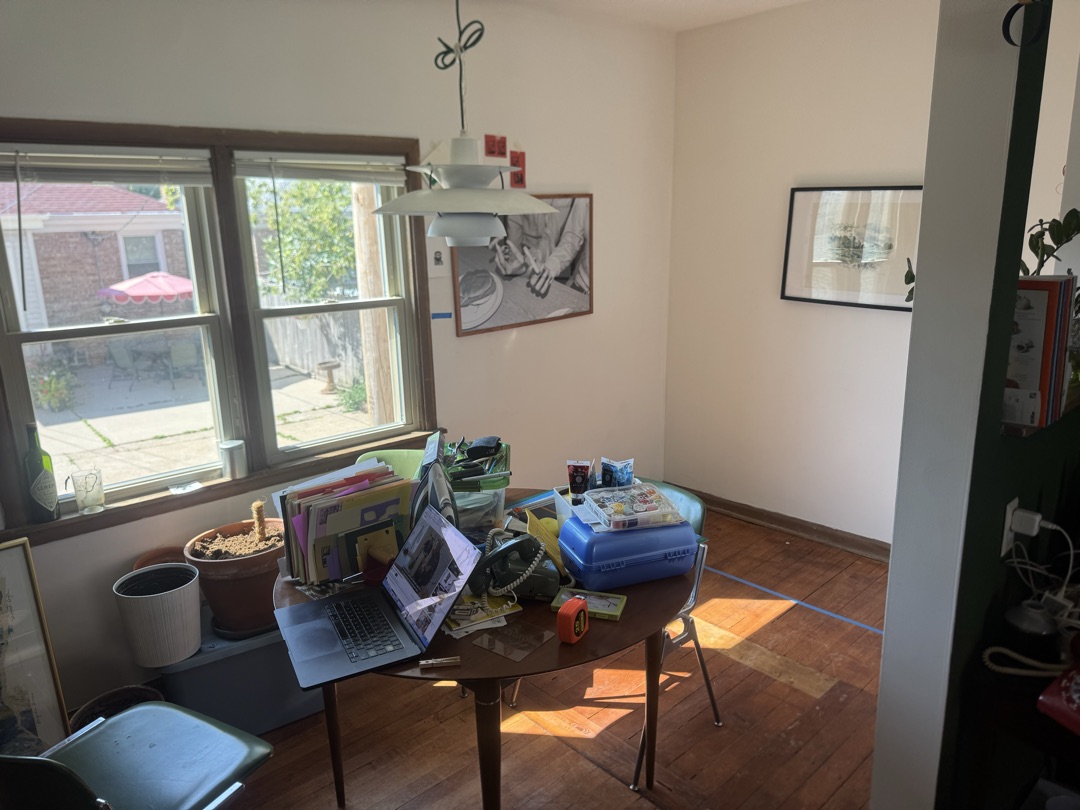 The dining room from the doorway: a round wooden table under a white pendant lamp, windows onto the back patio, and a line of blue painter's tape running across the floorboards
